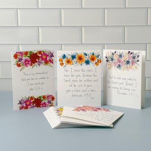May include: Three white greeting cards with floral watercolor illustrations and handwritten scripture verses. The left card features pink and red flowers with John 15:12. The center card has orange and blue flowers with Jeremiah 29:11. The right card shows pastel flowers with Revelation 21:5.