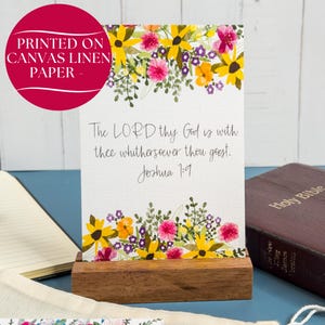 May include: A rectangular canvas linen paper print featuring a floral border of yellow, pink, and purple flowers. The text reads: The LORD thy God is with thee whithersoever thou goest. Joshua 1:9. The print is held upright by a rectangular wooden stand.