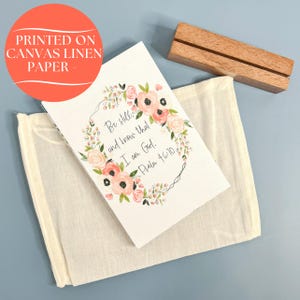 May include: A white card with a floral wreath design and the text "Be still and know that I am God. Psalm 46:10". The card is printed on canvas linen paper and is displayed on a wooden easel.