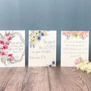 May include: Three rectangular art prints with floral designs and inspirational quotes. The first print features pink flowers and the text "Every good gift and every perfect gift is from above. James 1:17." The second print has blue flowers and the text "The joy of the LORD is your strength. Nehemiah 8:10." The third print has pink and orange flowers and the text "For I know the thoughts that I think toward you, saith the LORD, thoughts of peace, and not of evil, to give you an expected end. Jeremiah 29:11."