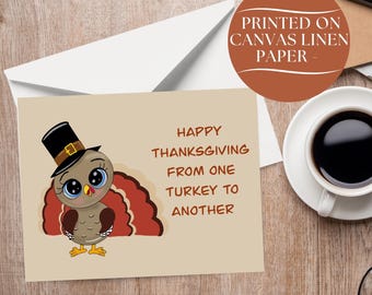 May include: A greeting card with a beige background featuring a cartoon turkey wearing a black top hat with a gold buckle. The text reads: HAPPY THANKSGIVING FROM ONE TURKEY TO ANOTHER. The card is printed on canvas linen paper and sits on a wooden surface.