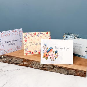May include: A collection of greeting cards with floral and bird designs. The cards feature phrases like "Holding you close in prayer," "I'm here for," and "Thinking of you." The cards are displayed on a wooden surface.