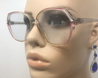 May include: Clear-framed eyeglasses with pink accents. The glasses have a large, square shape and are displayed on a mannequin. A pair of blue and silver drop earrings are also visible.