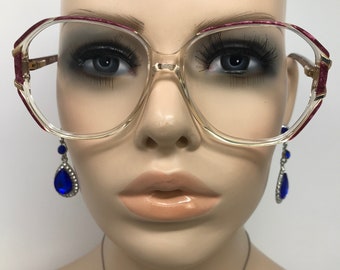 May include: Clear-framed eyeglasses with burgundy accents on the upper rims and temples. The glasses have a rectangular shape and are paired with blue teardrop earrings. The mannequin's skin tone is light.