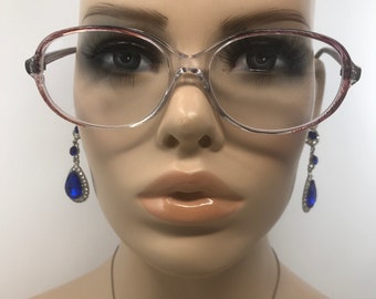 May include: Clear eyeglasses with a light brown rim and a clear bridge. The model is wearing blue teardrop earrings with silver accents. The image is a close-up of the face.