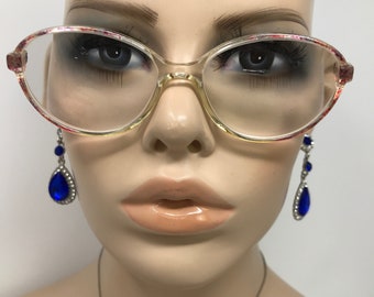Könnte beinhalten: Transparente ovale Brille mit floralem Muster an den oberen Rändern. Die Brille befindet sich auf einem Schaufensterpuppenkopf, mit blauen tropfenförmigen Ohrringen auf beiden Seiten.