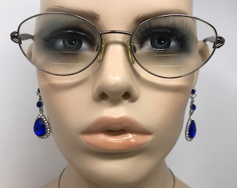 Peut inclure: Gros plan d'un mannequin portant des lunettes ovales avec une fine monture en métal marron. Le mannequin porte également de longs cils, des lèvres pulpeuses et des boucles d'oreilles pendantes bleues en forme de goutte d'eau avec des accents argentés.