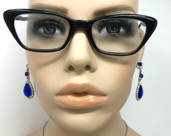 May include: Black cat-eye style eyeglasses with clear lenses are worn by a mannequin. The mannequin also wears dangling earrings with blue teardrop-shaped gems and clear rhinestones. The mannequin has full lips and long eyelashes.