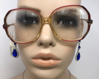 Könnte beinhalten: Übergroße Brille mit klaren Gläsern und einem hellbraunen Rahmen, akzentuiert mit roter Umrandung. Die Brille wird auf einer Schaufensterpuppe präsentiert. Ein Paar blaue Tropfenohrringe mit silbernen Akzenten.