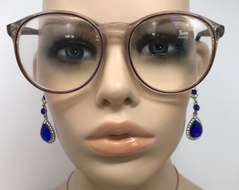 May include: Round-framed spectacles with a clear lens and a light brown tint. The glasses are worn with blue teardrop earrings with silver detailing. The image is a close-up of a mannequin head.