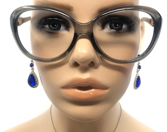 May include: Oversized gray eyeglasses with a cat-eye shape. The glasses are paired with blue teardrop earrings with silver accents and a small heart pendant necklace. The mannequin's skin tone is light.