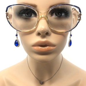 May include: Clear-framed eyeglasses with blue accents and gold-colored detailing. The glasses are paired with dangling blue teardrop earrings. A silver necklace with a heart pendant is also visible.