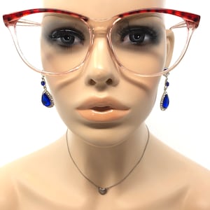 May include: Clear-framed eyeglasses with red and brown patterned upper rims. The mannequin wears blue teardrop earrings with silver accents and a small silver heart pendant necklace. The glasses are a cat-eye style.