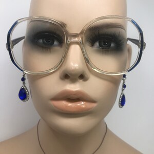 Op de afbeelding: Transparante oversized bril met blauwe accenten op de monturen. De bril wordt gecombineerd met blauwe druppelvormige oorbellen. De afbeelding toont een mannequinhoofd.