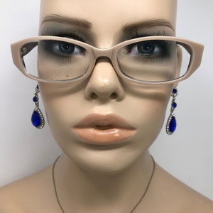 May include: A mannequin head wearing eyeglasses and jewellery. The eyeglasses have a light brown frame. The mannequin is wearing blue teardrop earrings with clear rhinestones. A silver necklace is also visible.