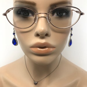 May include: Rose gold oval eyeglasses with clear lenses are worn by a mannequin. The mannequin also wears blue teardrop earrings and a silver heart pendant necklace. The glasses have a thin frame.