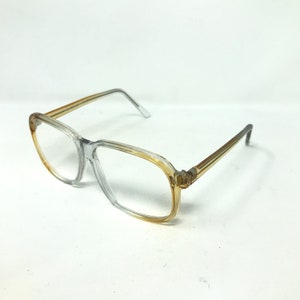 Puede incluir: Gafas con montura transparente con un tinte ámbar degradado en los bordes inferiores y exteriores de las monturas y las patillas. Las lentes son transparentes y el diseño general es rectangular. Las gafas están sobre un fondo blanco.