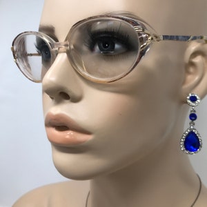 May include: Vintage eyeglasses with a light brown frame and clear lenses. The glasses have a rounded oval shape. A pair of blue and silver drop earrings are visible on the right side of the image.