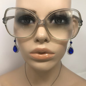 May include: Clear oversized eyeglasses with a light frame. The glasses are displayed on a mannequin with long eyelashes and a pair of blue teardrop earrings with silver accents.