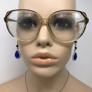 May include: Oversized eyeglasses with a light brown frame and black accents. The lenses are clear. The top corners of the frames have a red and white stripe design. The mannequin is also wearing blue teardrop earrings.