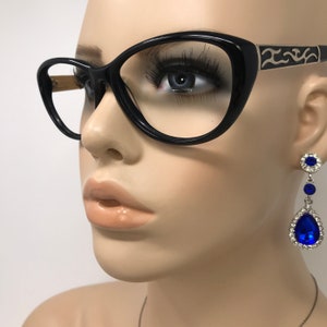 May include: Black cat-eye glasses with silver detailing on the arms. The image also shows long, dangling earrings with blue teardrop-shaped gems, surrounded by smaller clear crystals, and a round top. The earrings are silver-toned.