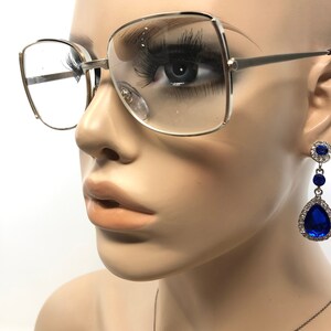 May include: Silver-framed eyeglasses with clear lenses and a square shape. The image also shows a pair of dangling earrings with a blue teardrop-shaped gem and smaller blue gems. The earrings are set in a silver metal.