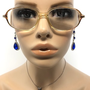 May include: Vintage eyeglasses with a light brown frame and tinted lenses. The glasses are paired with dangling blue teardrop earrings with silver accents. A silver necklace with a small heart pendant is also visible.
