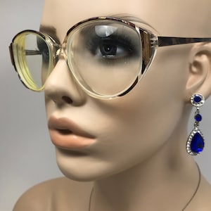 May include: Clear eyeglasses with a light brown and patterned frame. The glasses are on a mannequin. Dangling blue teardrop earrings with silver accents are also visible.