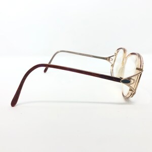 K&ouml;nnte beinhalten: Vintage Brille mit hellbraunem Rahmen und klaren Gl&auml;sern. Die B&uuml;gel sind dunkelbraun und der Steg ist silberfarben. Die Brille hat eine einzigartige, &uuml;bergro&szlig;e Form und ein kleines, dunkles ovales Detail am Rahmen.