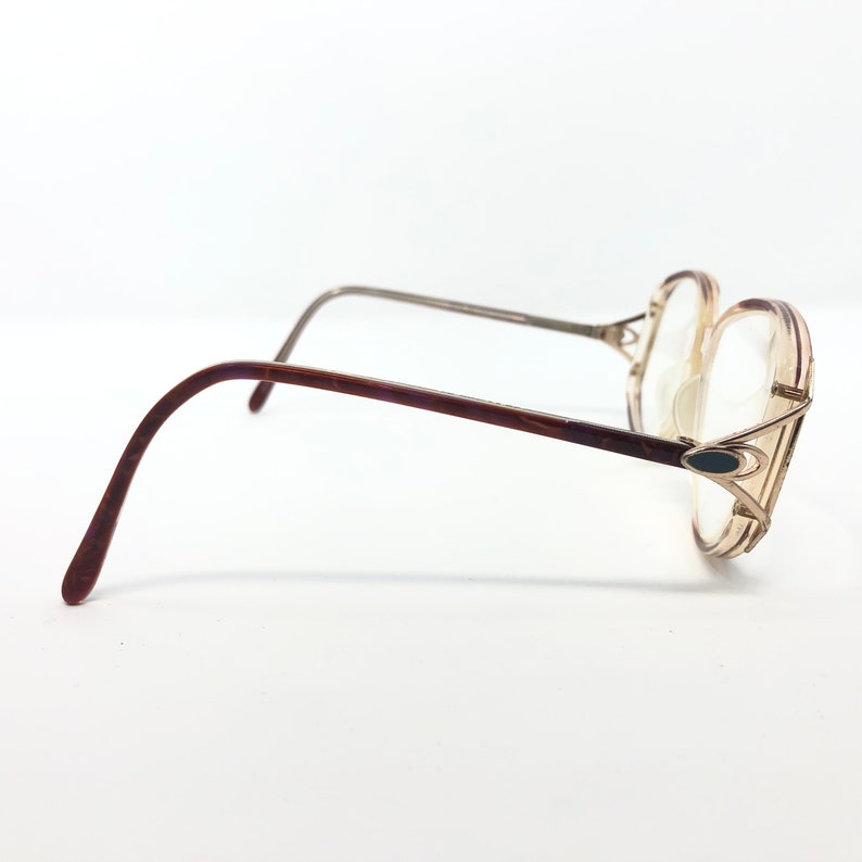 K&ouml;nnte beinhalten: Vintage Brille mit hellbraunem Rahmen und klaren Gl&auml;sern. Die B&uuml;gel sind dunkelbraun und der Steg ist silberfarben. Die Brille hat eine einzigartige, &uuml;bergro&szlig;e Form und ein kleines, dunkles ovales Detail am Rahmen.