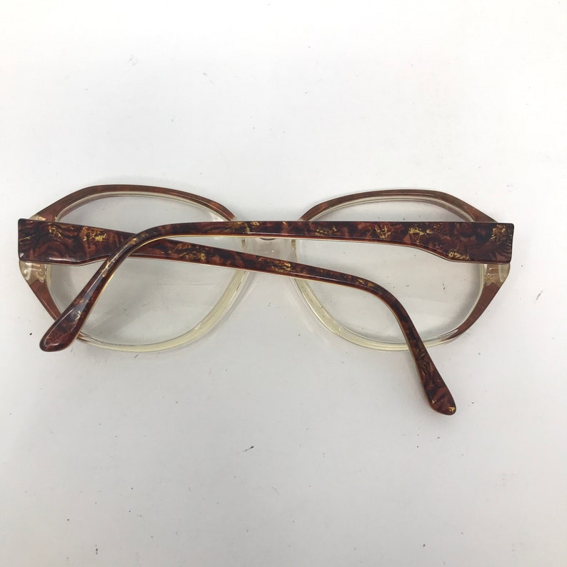 May include: Vintage eyeglasses with clear lenses and a brown and gold patterned frame. The glasses have a classic oval shape and a transparent lower rim. The arms of the glasses are also brown and gold.