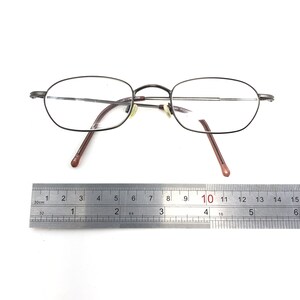 Puede incluir: Gafas de forma ovalada con una montura fina de metal oscuro y lentes transparentes. Las patillas son de color marr&oacute;n rojizo. Una regla met&aacute;lica debajo muestra medidas en cent&iacute;metros.