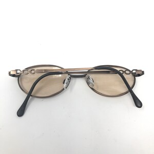 May include: Oval-shaped sunglasses with a bronze-colored frame and tinted lenses. The temples are black and the nose pads are clear. The frame features a chain-link design on the temples.
