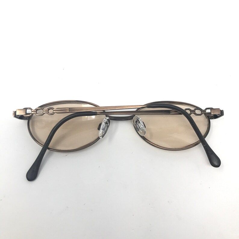 May include: Oval-shaped sunglasses with a bronze-colored frame and tinted lenses. The temples are black and the nose pads are clear. The frame features a chain-link design on the temples.