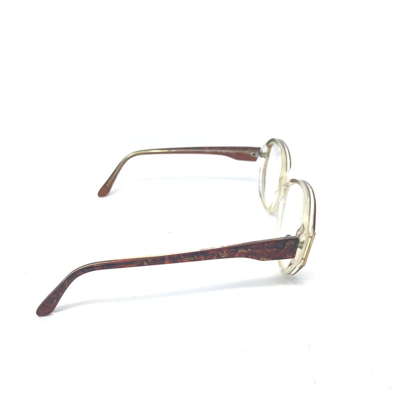 May include: Clear-framed eyeglasses with brown patterned temples and arms. The lenses are clear, and the frames have a rounded shape. The arms and temples have a brown, mottled pattern. The glasses are set against a white background.