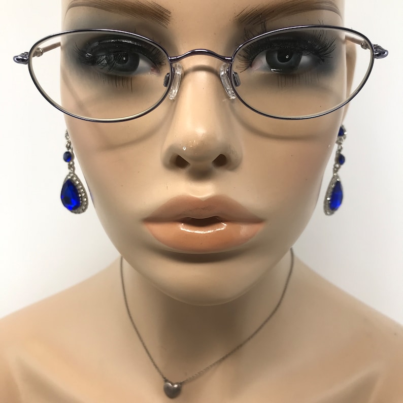 May include: Oval-shaped eyeglasses with a thin, dark frame. The image also features a pair of dangling earrings with large, blue teardrop-shaped gems and a small silver heart pendant necklace. The mannequin's face is visible.