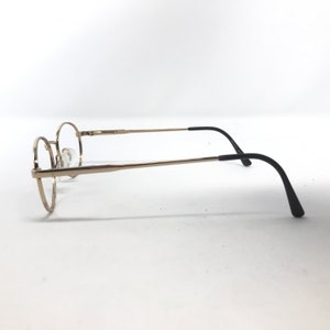 Pu&ograve; includere: Occhiali con montatura rotonda con montatura in metallo color oro e terminali neri. Le lenti sono trasparenti e gli occhiali sono su uno sfondo bianco. Il design &egrave; semplice e classico.