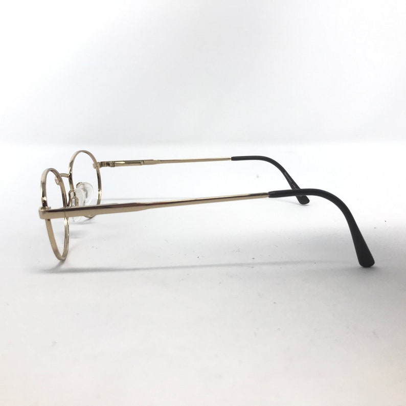 Pu&ograve; includere: Occhiali con montatura rotonda con montatura in metallo color oro e terminali neri. Le lenti sono trasparenti e gli occhiali sono su uno sfondo bianco. Il design &egrave; semplice e classico.