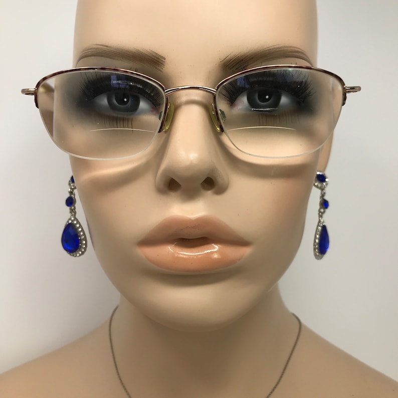 May include: Gold-rimmed eyeglasses with a half-frame design. The glasses have clear lenses and are set on a mannequin head. The mannequin is also wearing blue teardrop earrings with silver accents.