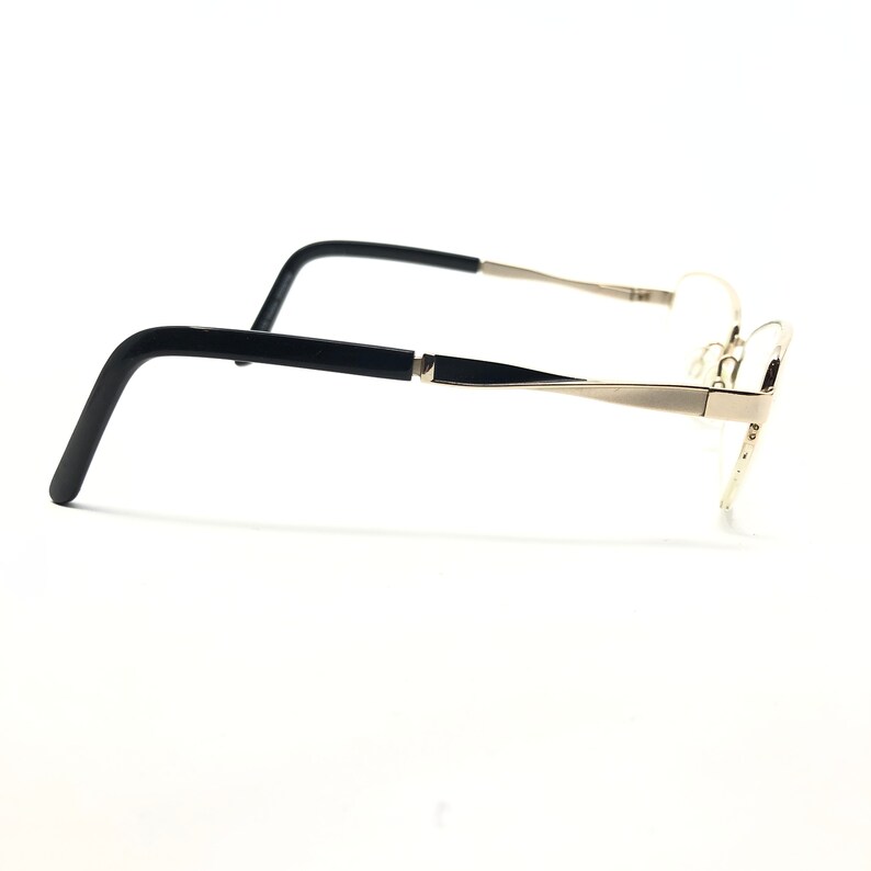 Pu&ograve; includere: Un paio di occhiali con montatura in metallo color oro e aste nere. Gli occhiali sono mostrati di profilo su sfondo bianco. Le aste hanno una forma rettangolare e sono collegate alla montatura con una piccola cerniera.