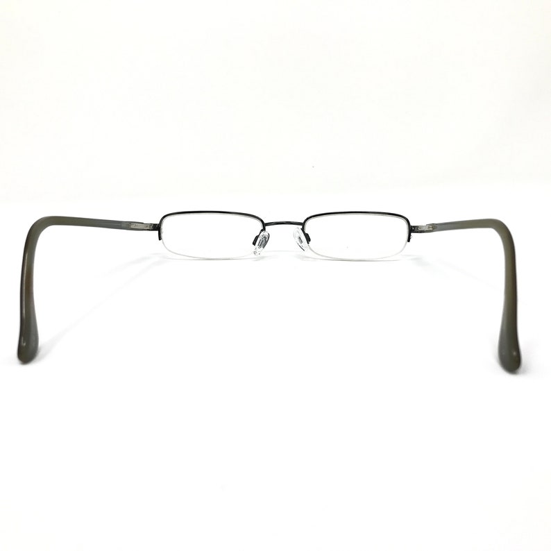 K&ouml;nnte beinhalten: Eine Brille mit einem dunkelgrauen Metallrahmen und klaren Gl&auml;sern. Die Rahmen haben ein Halbrand-Design, wobei die Gl&auml;ser durch einen d&uuml;nnen Draht am unteren Rand gehalten werden. Die B&uuml;gel sind dunkelgrau.