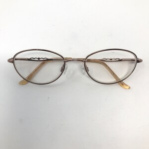 May include: Oval-shaped eyeglasses with a thin, bronze-colored metal frame. The lenses are clear, and the temples have a decorative wave-like design. The nose pads are transparent, and the earpieces are a light beige color.