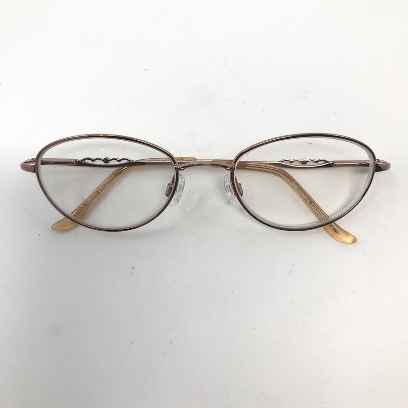 May include: Oval-shaped eyeglasses with a thin, bronze-colored metal frame. The lenses are clear, and the temples have a decorative wave-like design. The nose pads are transparent, and the earpieces are a light beige color.