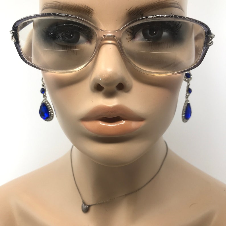 May include: Clear-framed eyeglasses with a patterned top edge are worn by a mannequin. The mannequin also wears dangling blue teardrop earrings and a silver necklace with a small pendant. The background is white.