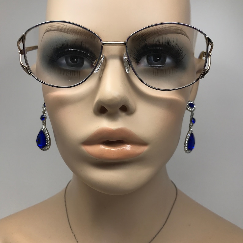 Op de afbeelding: Goudkleurige bril met een cat-eye vorm. De bril heeft heldere glazen en wordt getoond op een mannequin. Blauwe druppelvormige oorbellen met zilveren accenten zijn ook zichtbaar.