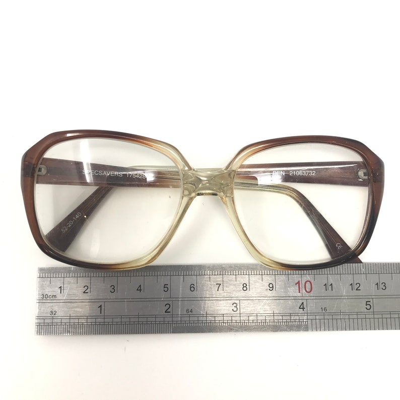May include: Brown and clear eyeglasses with a rectangular shape. The glasses have a gradient tint and are measured with a ruler. The frame has the text "SPECSAVERS 175428" and "DEN 21063732".