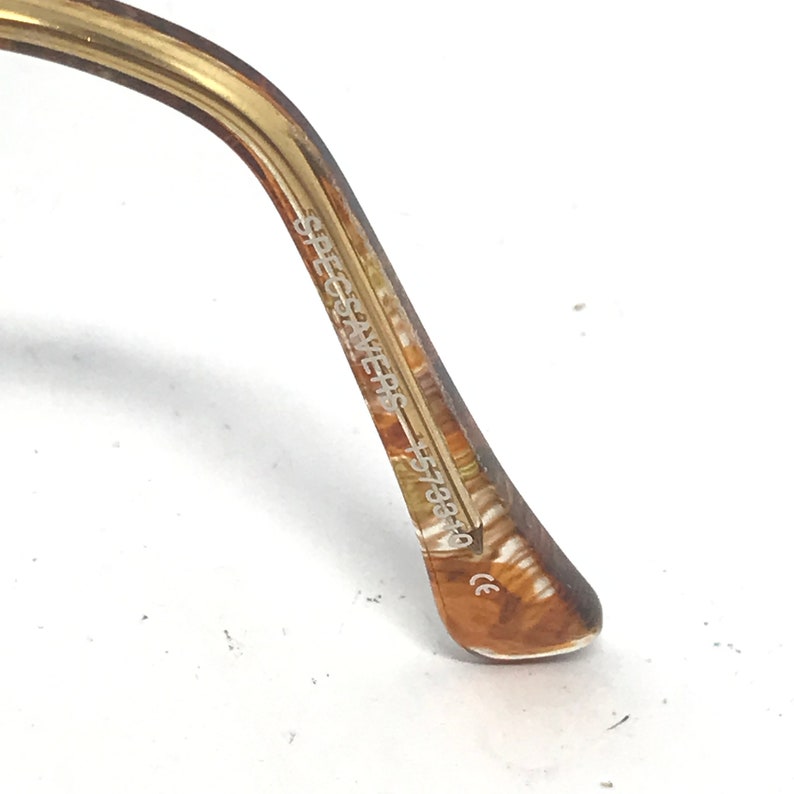 May include: Close-up of the arm of eyeglasses. The arm is a marbled brown and tan color. The text "SPECSAVERS" and "5738F0" is printed on the arm. The end of the arm has a small "CE" marking.