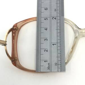 May include: A pair of eyeglasses with a clear frame and a brown-tinted upper frame. A metal ruler is placed over the lens, measuring approximately 2 inches in height. The glasses are set against a white background.