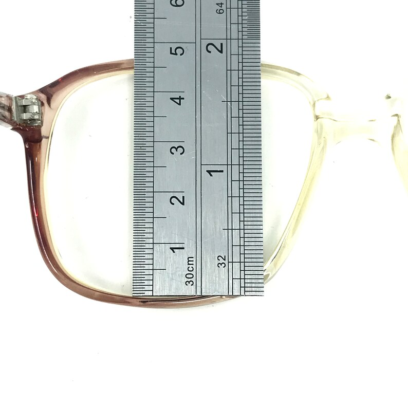 May include: Clear eyeglasses with a light brown frame are measured with a ruler. The ruler shows the glasses are approximately 2 inches wide. The glasses have a rectangular shape and are set against a white background.