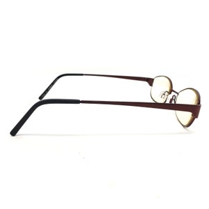 May include: A pair of eyeglasses with a maroon frame and black temples. The lenses are round and the frame has a minimalist design. The glasses are set against a white background.